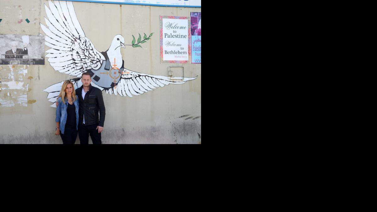 
Journalist Mariana van Zeller, left, and chef Michael Voltaggio, hosts of Travel Channel’s “Breaking Borders,” pose in front of an art piece showing a dove in a bulletproof vest by graffiti artist Banksy in Bethlehem, Palestine. “Breaking Borders” is a travelogue, cooking show and dining-table summit combined.
