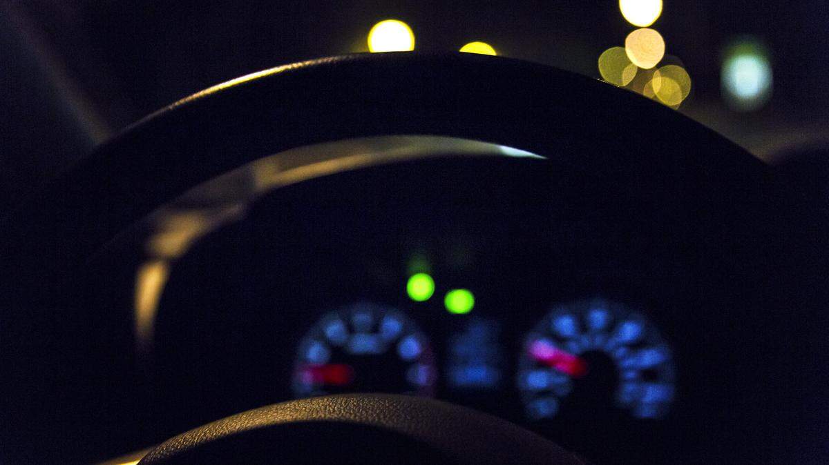 Low angle view through a car steering wheel at the illuminated dashboard and speedometer, with abstract, defocused multi colored lights up ahead in the night darkness.