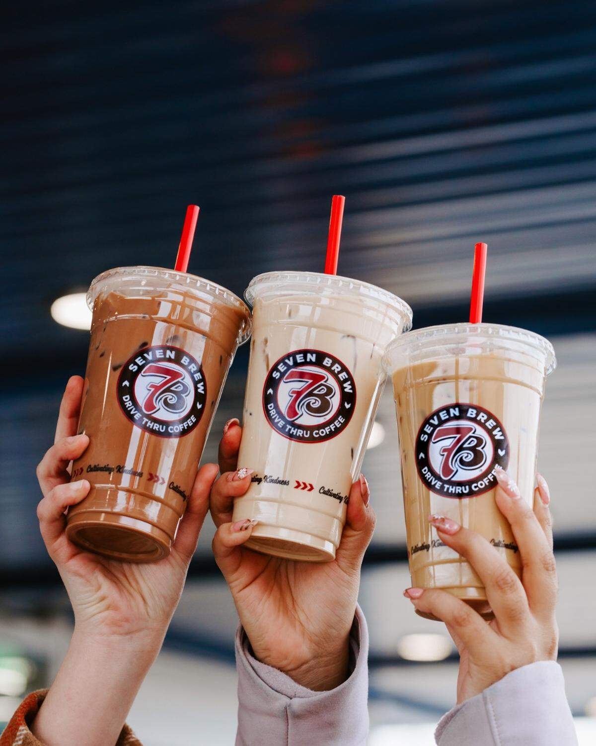 Three people are holding up clear plastic cups of iced coffee drinks with red straws. The cups are from the brand 7 Brew Coffee, and the drinks are different shades of brown, indicating various flavors or types of coffee.