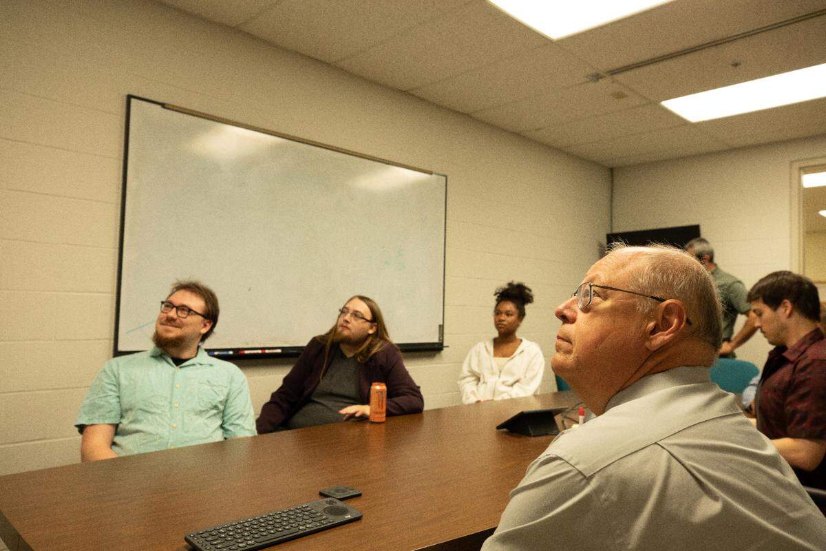 Five people sit around a table looking upwards towards a screen not-pictured. 