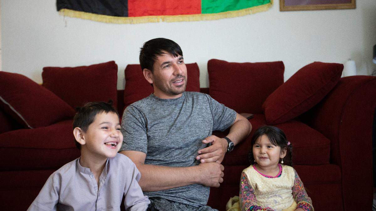 Shakil Alakozai sits under an Afghanistan flag in his living room with his 7-year-old son Shokaib and 5-year-pld daughter Halima. (May 21, 2022)
