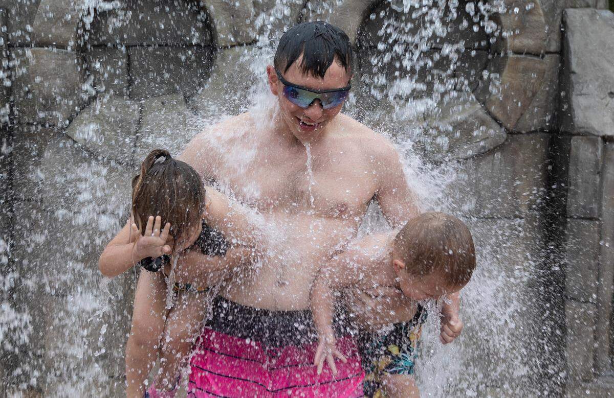 Tyler Fehl carries his two siblings, Lillian Lee, left, and Liam Lee, right, through a fountain at the splash pad at Evergreen Park recently. Three of the city’s splash pads opened early. The rest, plus city pools, are set to open on Memorial Day.
