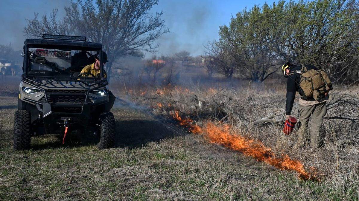 In this photo courtesy of DVIDS, U.S. Air Force Wildland Fire Branch wildland firefighter Cody Jolovich sprays water on cut grass while Alex Mermigkas, Wildland Fire Branch module leader, ignites tall grass during a controlled burn at McConnell Air Force Base on March 20, 2025.