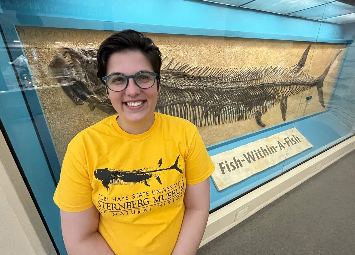 Aly Baumgartner stands in front of the Sternberg Museum’s famous fish-within-a-fish display, a fossil that’s also commemorated on her shirt.