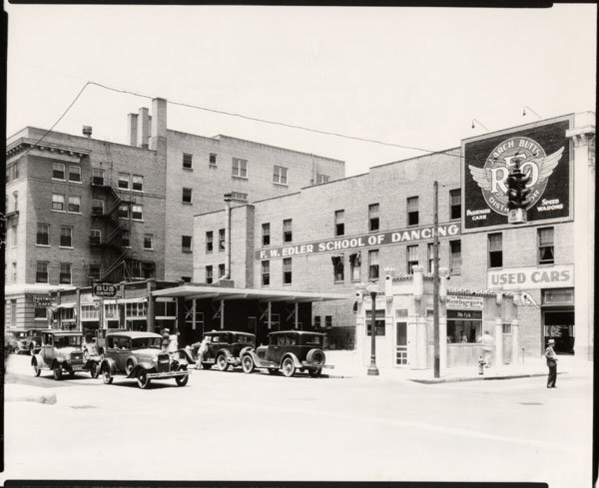 Looking northwest in 1931 from the intersection of First Street and Lawrence (now known as Broadway) in Wichita, Kansas. Businesses visible include: White Castle Hamburgers, Union Bus Depot, Tostwich Cafe, Butts New & Used Cars, Edler School of Dancing. The automobile in the left foreground appears to have an "Air Capital" emblem mounted on the grille.