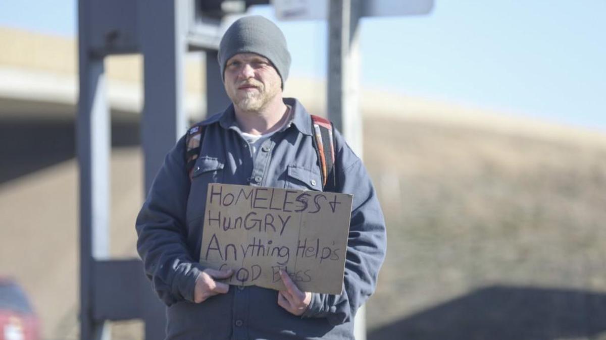 A man stands by the intersection of K-96 and Rock Road holding a sign as cars drive by.
